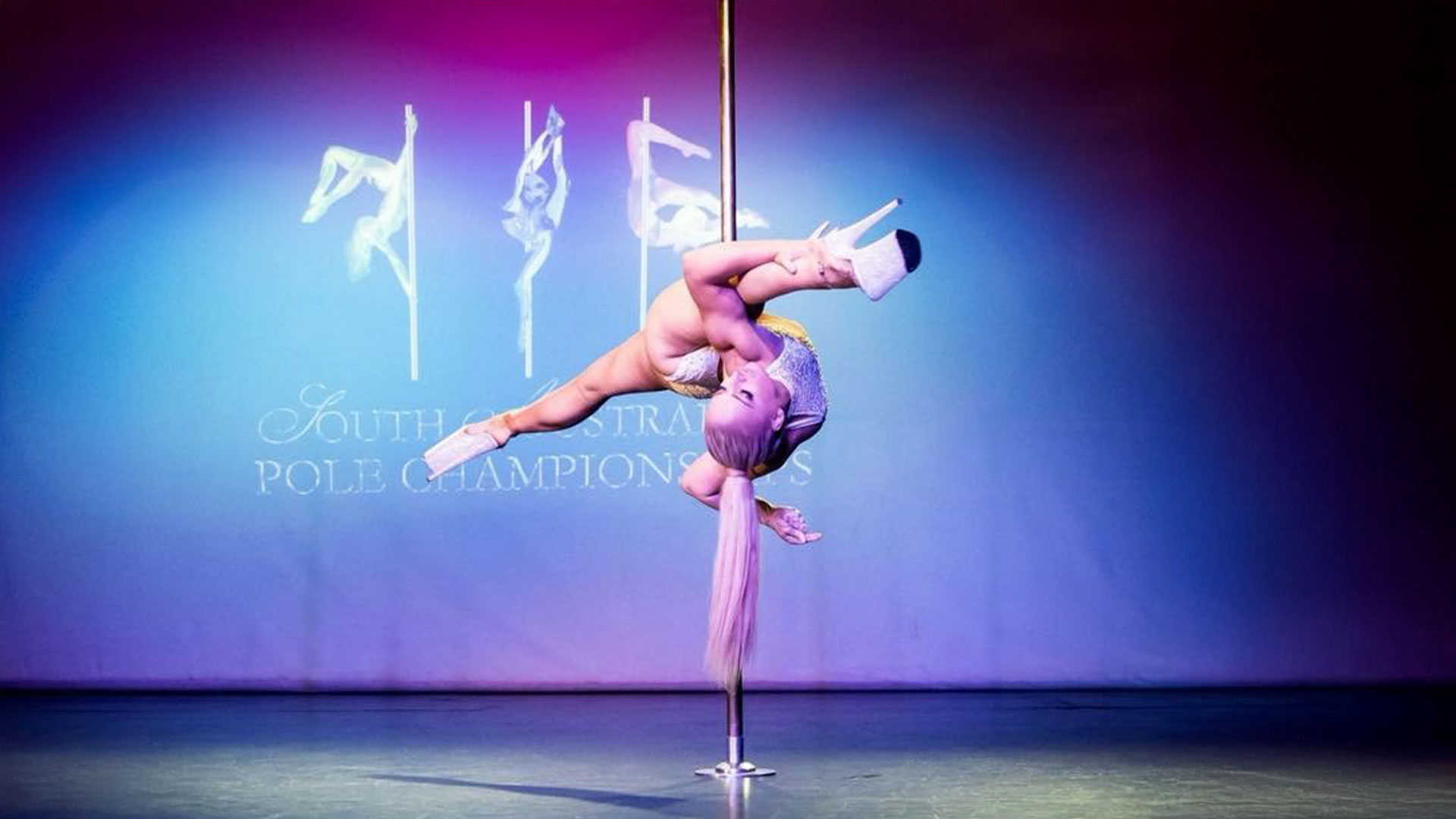 A pole dancer in a white performance outfit and high heels is performing a side split on a pole.